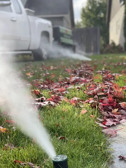 A sprinkler head blowing out air and mist across red autumn leaves on a lawn