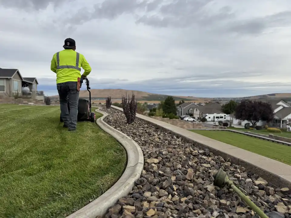 A First Choice crew member mowing along a long curved concrete curb and rock bed, valley hills behind